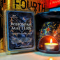 A wax melt package labeled 'Wingspan Matters' stands in front of a decorative wax melt burner with a lit tealight candle. The package shows a dark blue space-themed label with bat wings and the scents 'Cashmere,' 'Mahogany,' and 'Oudh.