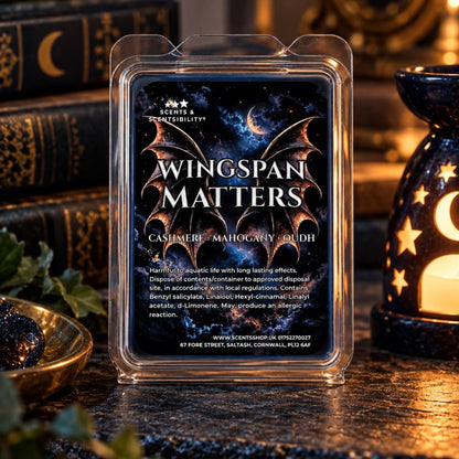 A clear plastic clamshell container holding a wax melt package labeled 'Wingspan Matters' stands on a dark tabletop. The package artwork shows a black bat wing over a starry blue and purple night sky.