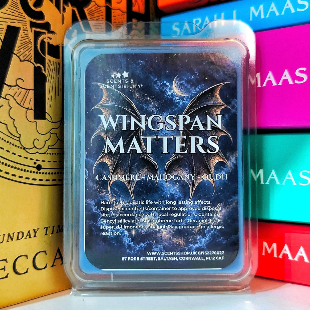 A scented wax melt in a clear plastic clamshell container with a blue label reading 'Wingspan Matters' and 'Cashmere + Mahogany +oudh.' The wax melt is photographed upright in front of colorful books, with one gold book called Fourth Wing partially visible on the left.