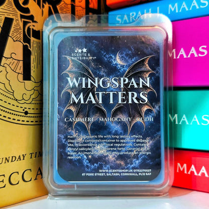 A scented wax melt in a clear plastic clamshell container with a blue label reading 'Wingspan Matters' and 'Cashmere + Mahogany +oudh.' The wax melt is photographed upright in front of colorful books, with one gold book called Fourth Wing partially visible on the left.