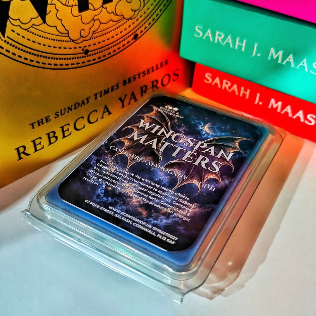 A scented wax melt, labeled 'Wingspan Matters', shown in a clear plastic clamshell package on a shelf books in the background.