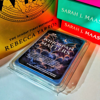 A scented wax melt, labeled 'Wingspan Matters', shown in a clear plastic clamshell package on a shelf books in the background.