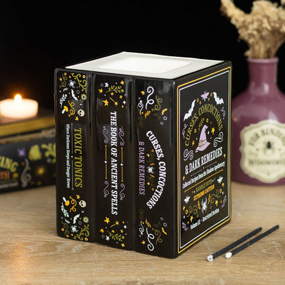 A black, decorative wax melt and oil burner, shaped like a stack of books about ancient spells and curses