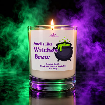 A lit scented candle in a clear glass jar with a white label reading 'Smells like Witches Brew' and a black cauldron graphic with green potion. The candle is photographed against a dark background with dramatic green and purple smoke effects.