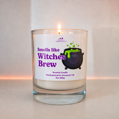 A lit scented candle in a clear glass jar with a white label that reads 'Smells like Witches Brew' in purple text, featuring a black cauldron with green potion illustration. The candle is photographed against a soft beige background with a warm glow near the base.