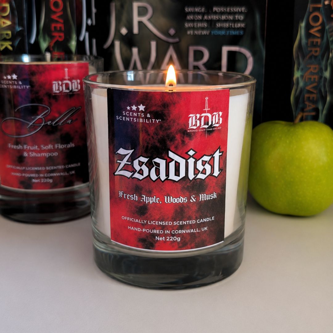 Candle with a red label featuring the word 'Zsadist' in front of J.R. Ward books and a green apple.