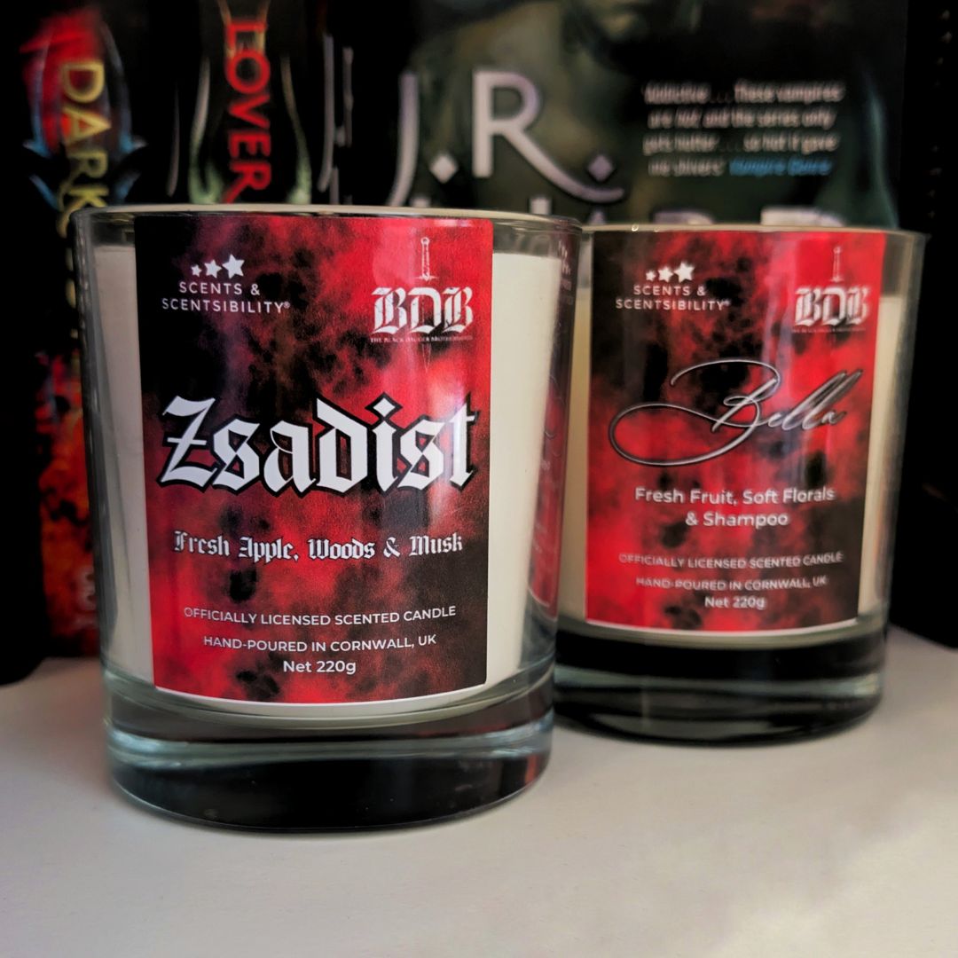 Two scented candles labeled 'Zsadist', and 'Bella' on a white surface with books in the background.